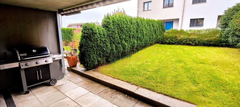 Well-maintained garden with a neatly trimmed hedge, a small lawn, and a covered patio area featuring a grill and a potted plant.