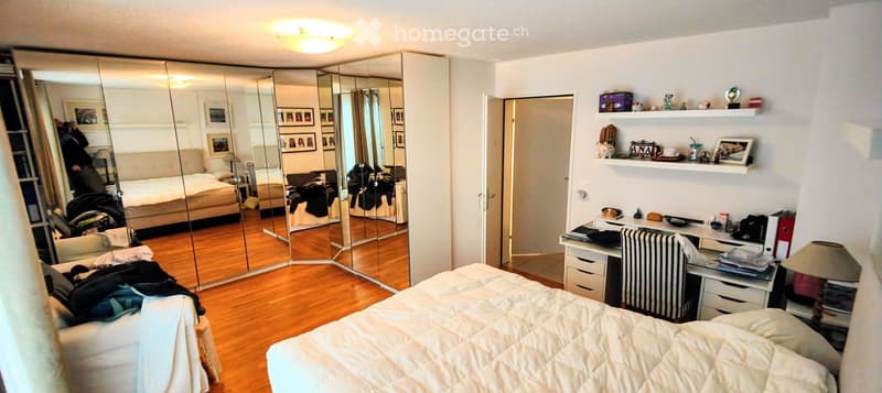 Well-lit bedroom with wooden flooring, large mirror, bed, desk, and shelves with decorative items.