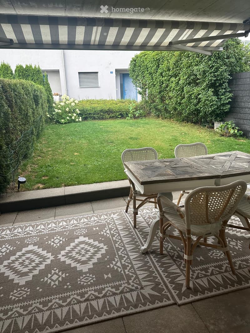 Outdoor patio with a wooden table, wicker chairs, patterned rug, and a well-maintained lawn.