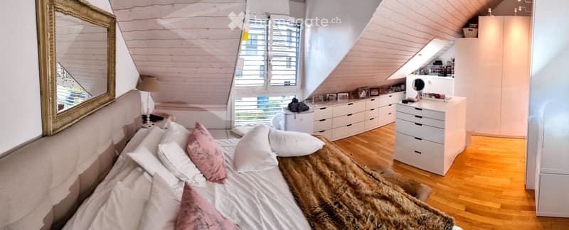 Cozy attic bedroom with wooden slanted ceilings, large windows, a neatly made bed with pillows and a fur throw, a large mirror, a lamp, and built-in storage including a wardrobe and drawer unit.