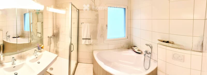Modern bathroom with a large mirror, double sink, bathtub, and a glass-enclosed shower. White tiles and a window provide natural light.