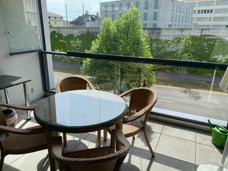 Balcony with a round glass table and four wicker chairs, overlooking a cityscape with trees and buildings.