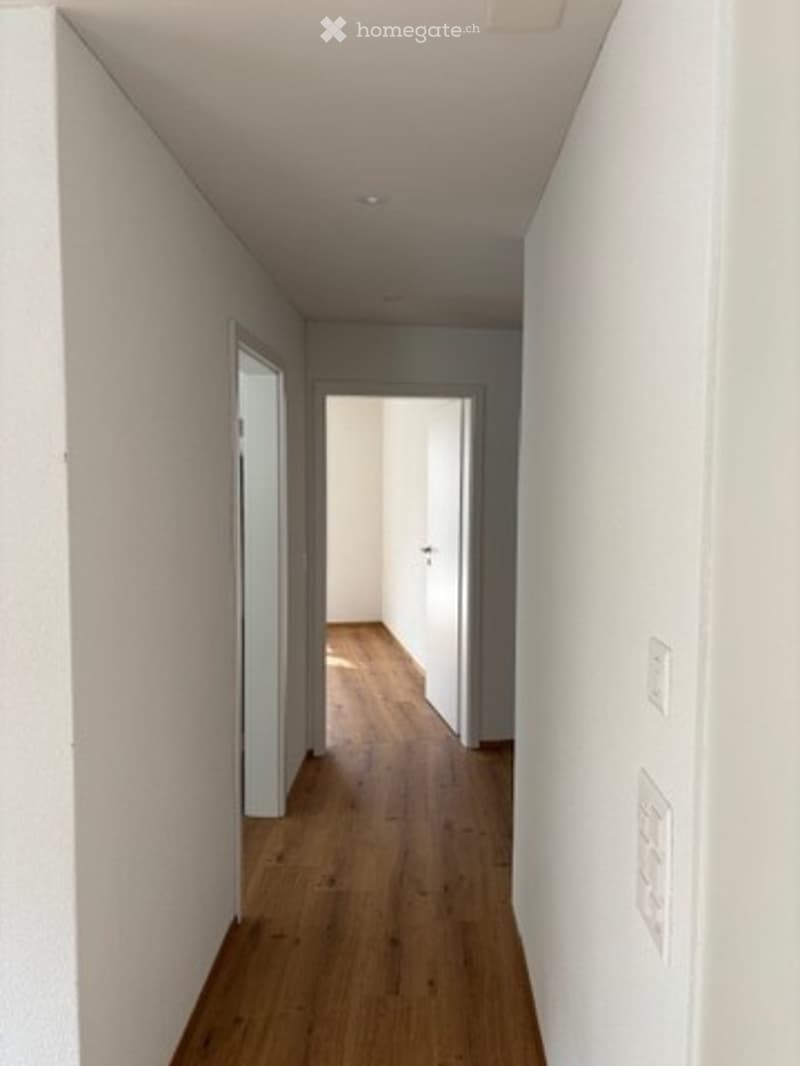 A clean, modern hallway with light wooden flooring, white walls, and recessed ceiling lighting. The hallway features a partially open door on the left and an open doorway at the end leading to another room.