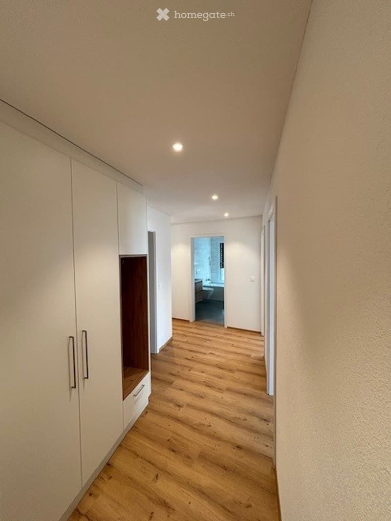 Empty hallway with light wooden flooring, white walls, built-in white closet with mirrored doors, and recessed ceiling lights.