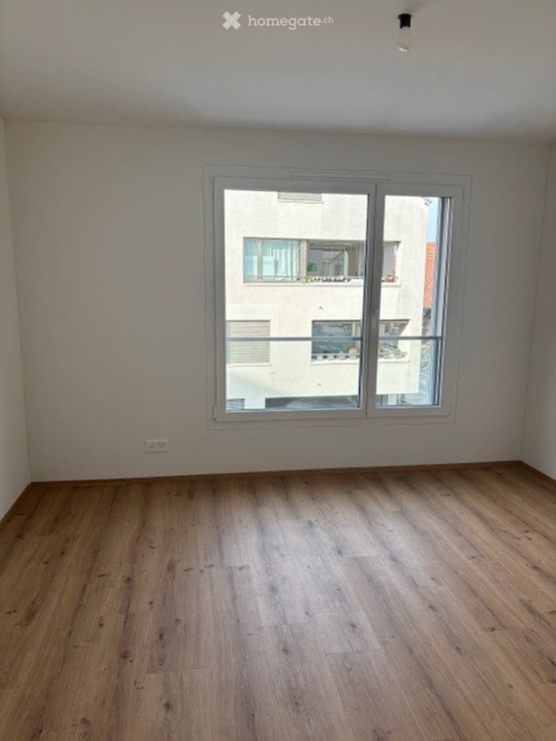 Empty room with wooden flooring and large window offering a view of a neighboring building. No furniture or equipment present.