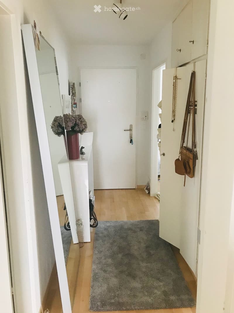 A narrow hallway with wooden flooring, a grey rug, white walls, and a closed door at the end. A small console table with a vase of flowers is visible on the left, and a coat rack with bags is on the right.
