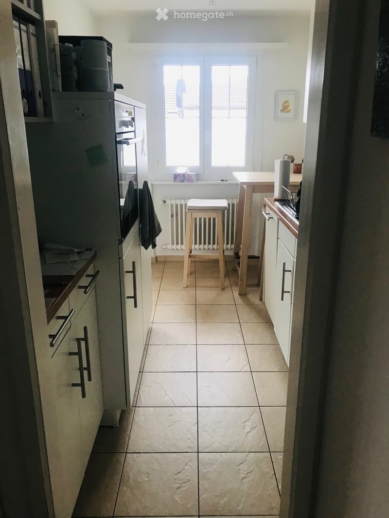 Modern kitchen with light-colored cabinets, a central island, a stool, and a refrigerator. The floor is tiled, and there is a window above the sink area.