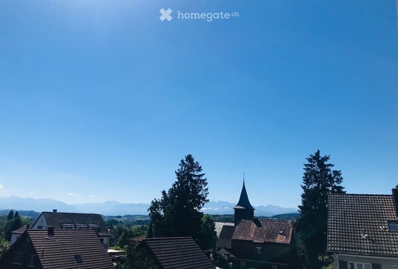 A scenic view of a village with traditional houses, a church tower, and surrounding mountains under a clear blue sky.