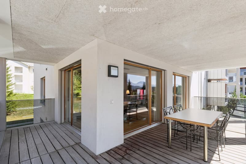 Modern balcony with wooden decking, white table, and chairs, large glass sliding doors leading to interior, scenic mountain view.