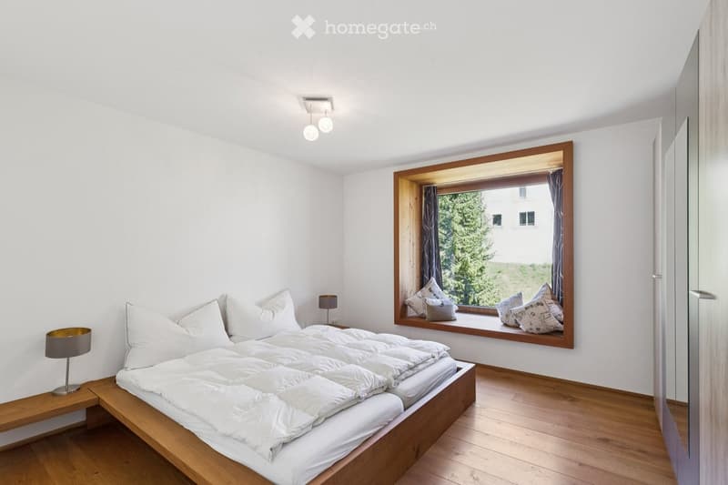 Modern bedroom with a large wooden bed, white bedding, nightstand with lamp, and a window seat with cushions.