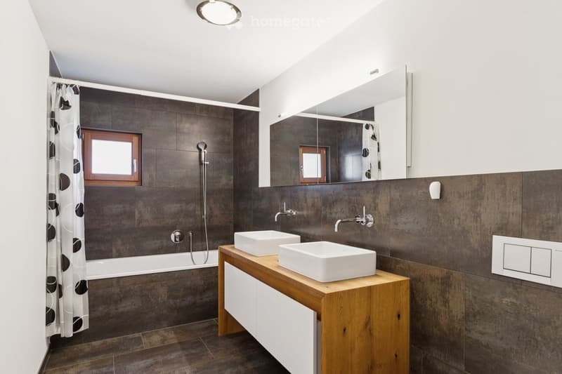 Modern bathroom with dark grey tiles, a wooden vanity with two white sinks, a bathtub with a shower curtain, and a large mirror above the vanity.