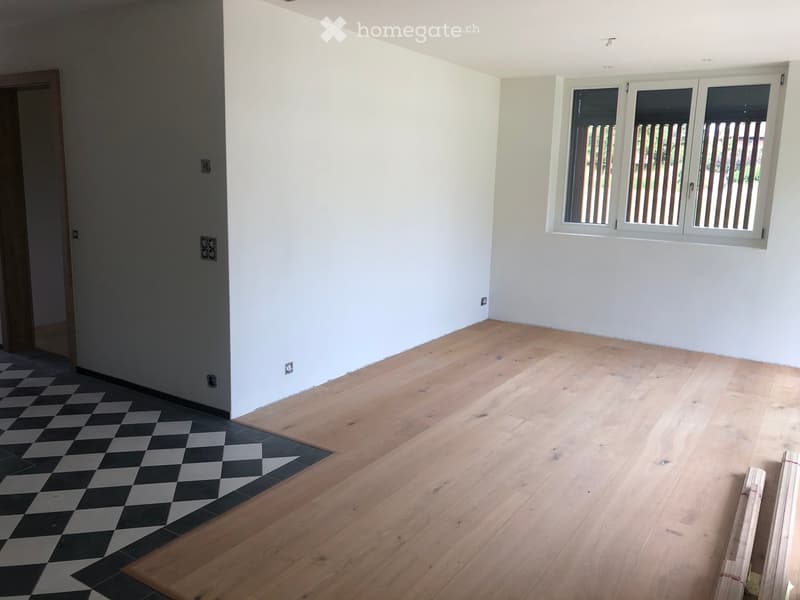 Empty room with light wooden flooring and white walls, featuring a checkered black and gray tile section near the doorway and a window with white frames and vertical blinds.
