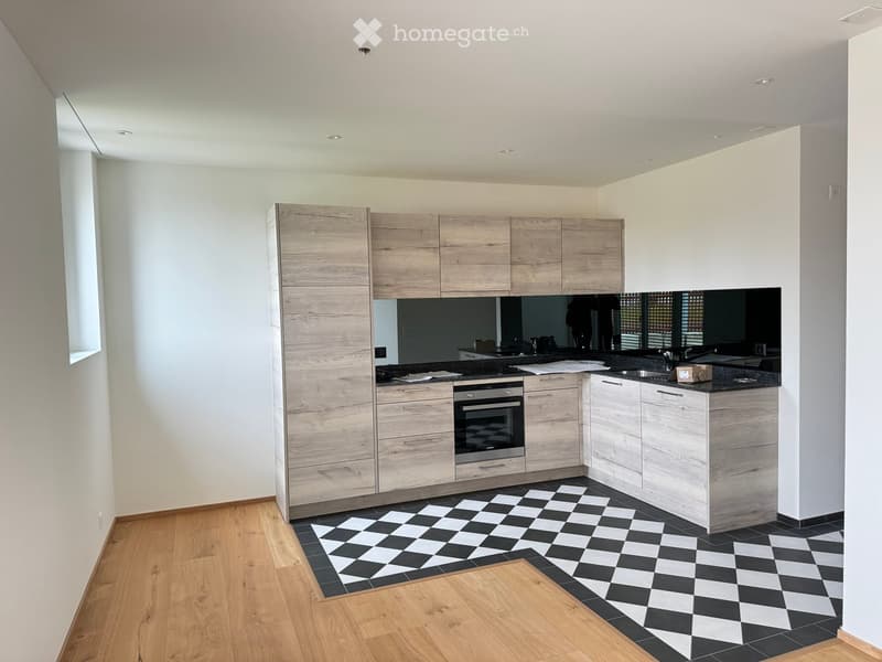 Modern kitchen with light wood cabinets, stainless steel oven, black countertop, and checkered black and white floor tiles.