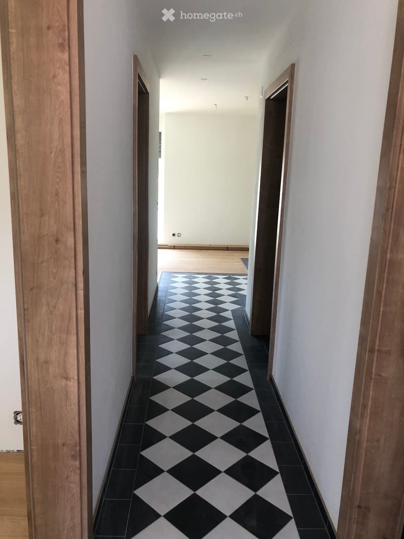 A long, narrow hallway with a black and white checkered tile floor, light-colored walls, and wooden doors on both sides.