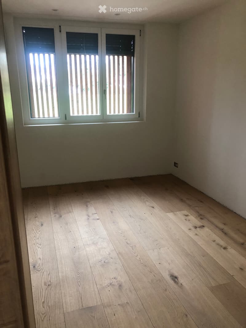 Empty room with light wooden flooring and white walls, featuring a set of three windows with white frames and vertical blinds.