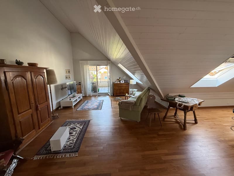Bright attic living space with wooden floors, slanted ceilings, large windows, and skylights. Features a sofa, coffee table, armchair, TV, and a wooden cabinet.