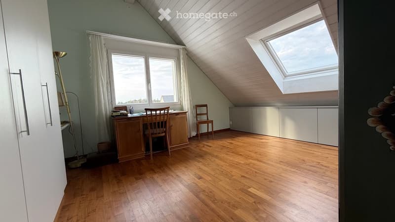 Bright attic room with wooden flooring, slanted ceiling, large windows, and skylight. Contains a wooden desk with a chair, a floor lamp, and a cabinet.