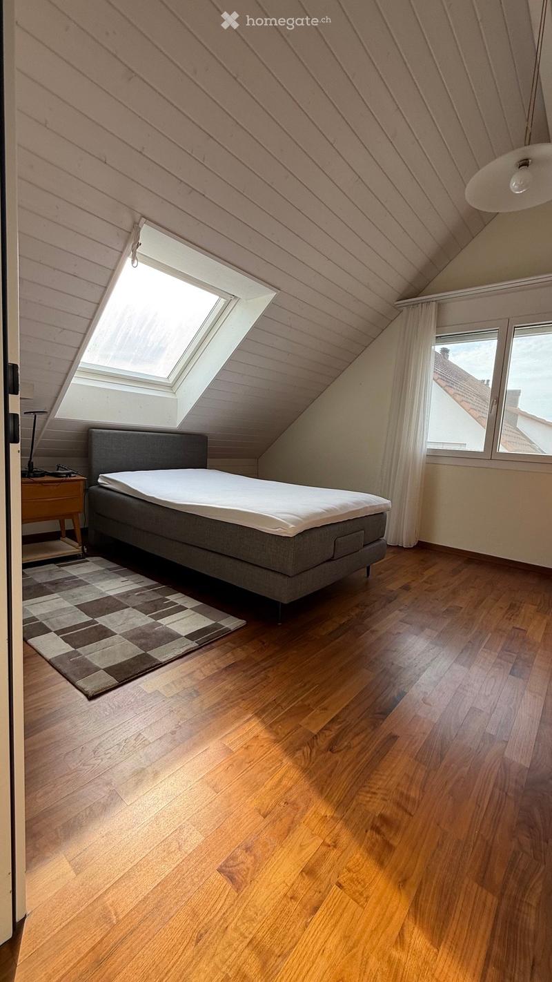 Cozy attic bedroom with wooden floors, slanted wooden ceiling, skylight window, and large window with view. Features a grey bed with white bedding, a patterned rug, and a nightstand with a lamp.