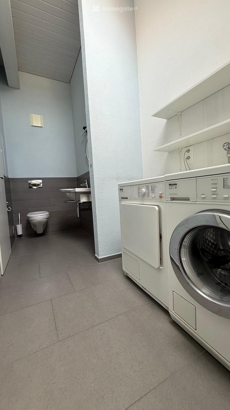 Modern bathroom with a toilet, sink, and washing machine. The space features light grey tiled walls and a grey tiled floor.