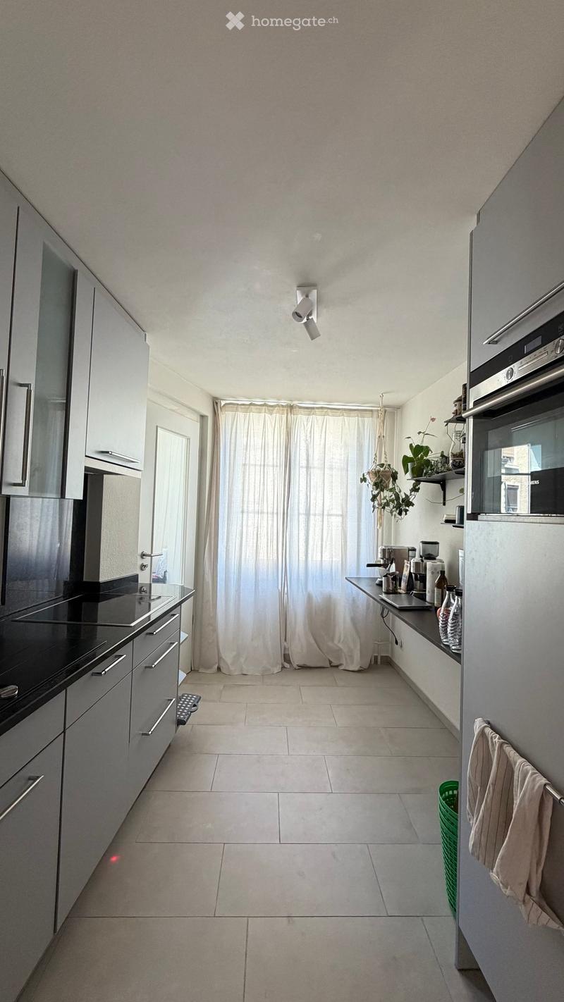 Modern kitchen with grey cabinets, white tiled floor, black countertop, sink, fridge, oven, coffee maker, laptop, and a potted plant on the windowsill.