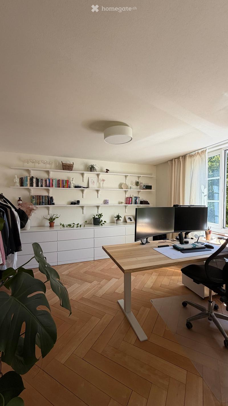 A modern, well-lit home office with a wooden desk, ergonomic chair, dual monitors, shelves filled with books and decorative items, and large windows with curtains.