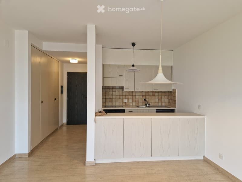 Modern kitchen with light wood cabinets, a central island, and pendant lighting. The space features a sleek design with a polished wooden floor and a visible doorway leading to another room.