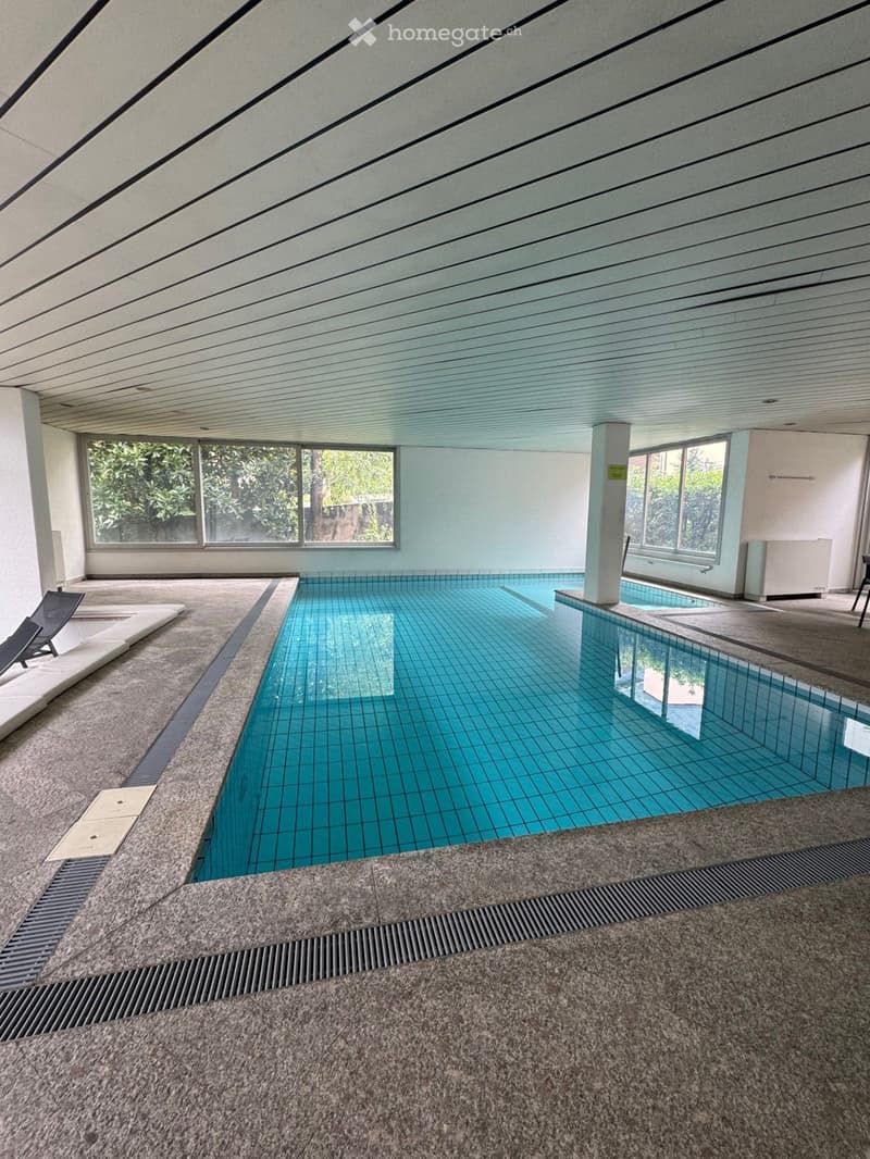 Indoor swimming pool area with large windows, tiled pool, lounge chairs, and a modern ceiling design.