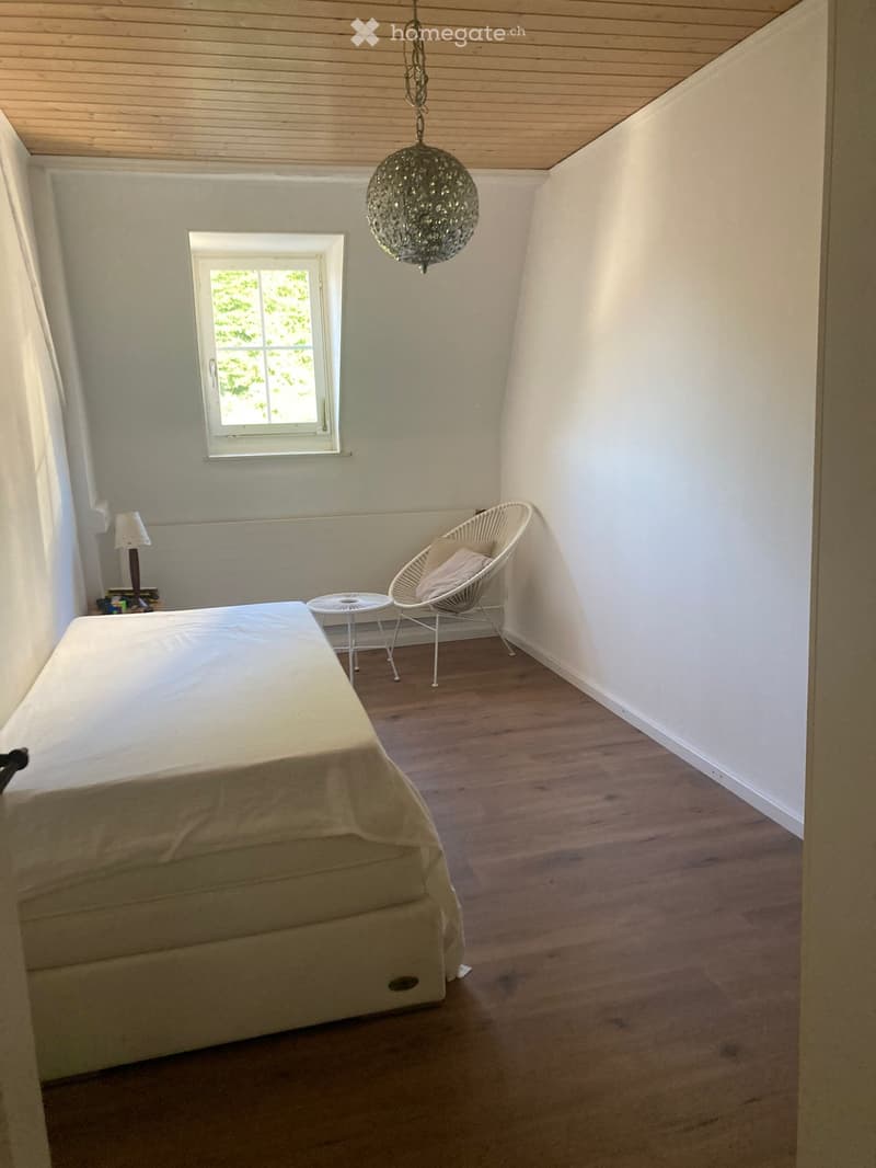 Cozy bedroom with wooden flooring, a bed, a hanging chandelier, a small round table, and a wicker chair.