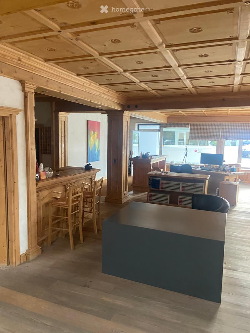 Office space with a reception desk, wooden stools, office desk, computer, and bookshelves. The ceiling features decorative wooden paneling.