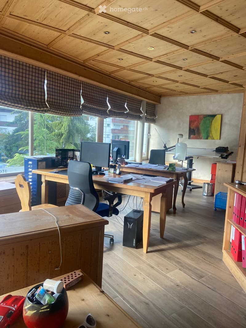 Office space with wooden flooring, large windows with blinds, two desks with computers, chairs, and various office supplies.