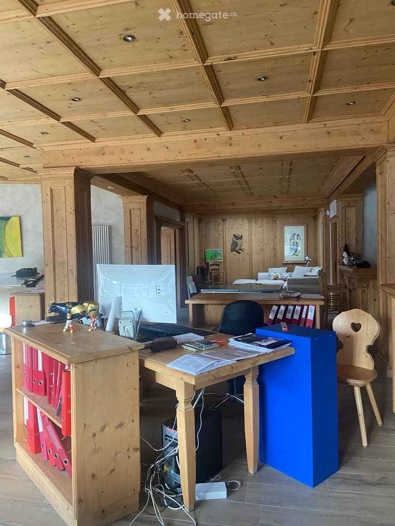 A spacious, well-lit office with wooden beams and a high ceiling. Features include a large wooden desk with a laptop, screen, and various office supplies, a comfortable chair, a filing cabinet, and a cozy sitting area with a sofa and a framed picture on t