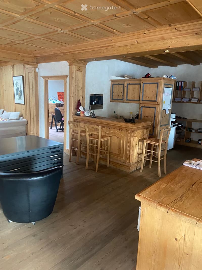 A cozy, wooden-paneled room with a central bar, bar stools, a leather chair, a sofa, and a small kitchen area with cabinets and a refrigerator.
