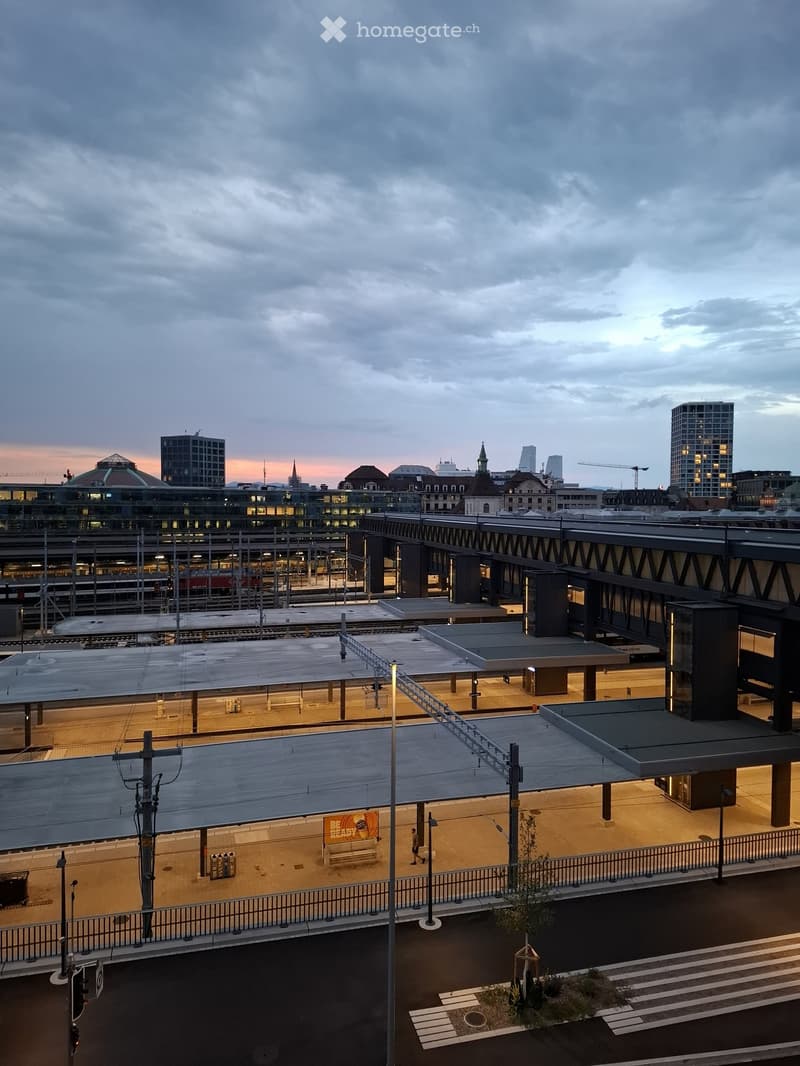 A train station platform with a bench, modern architecture, and a cityscape in the background.