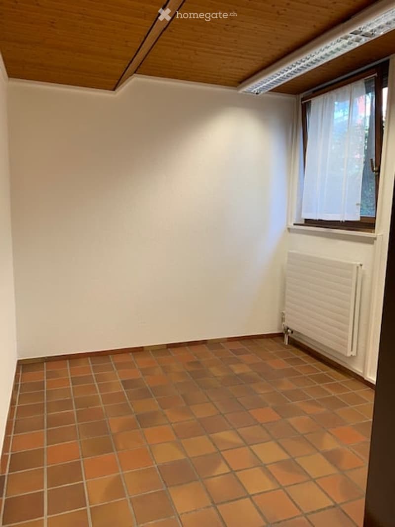 Empty room with white walls, wooden ceiling, and orange tiled floor. A window with white curtains and a white radiator are visible.