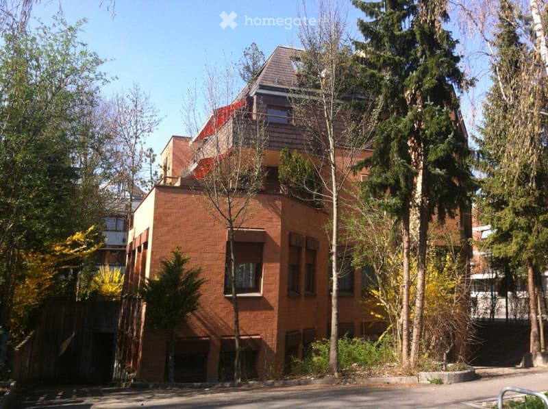 Modern two-story brick building with large windows and a sloped roof, surrounded by trees and greenery.