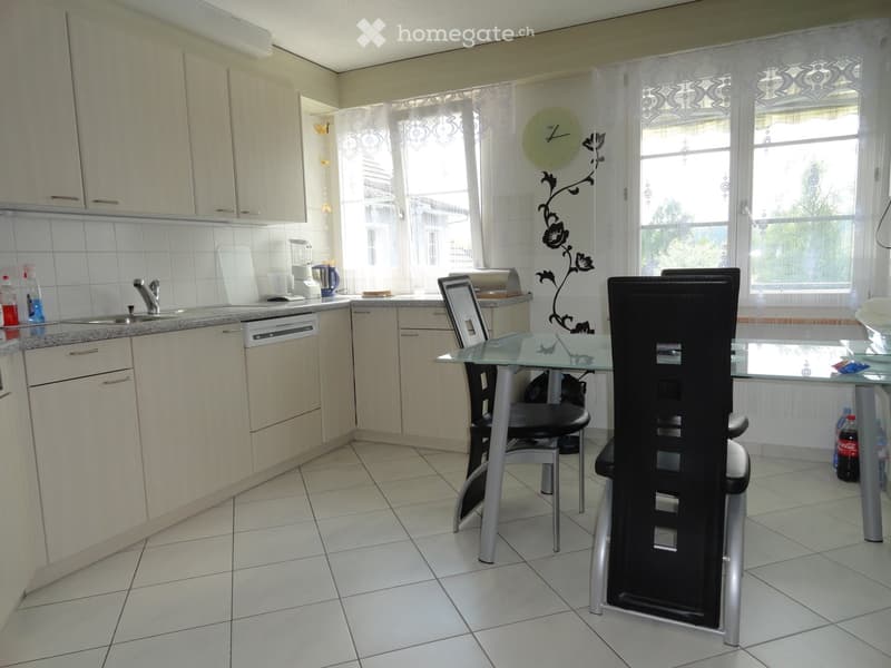 Bright kitchen with white tiled walls, light-colored cabinets, and a tiled floor. Features a sink, dishwasher, and a small dining area with a glass table and black chairs.