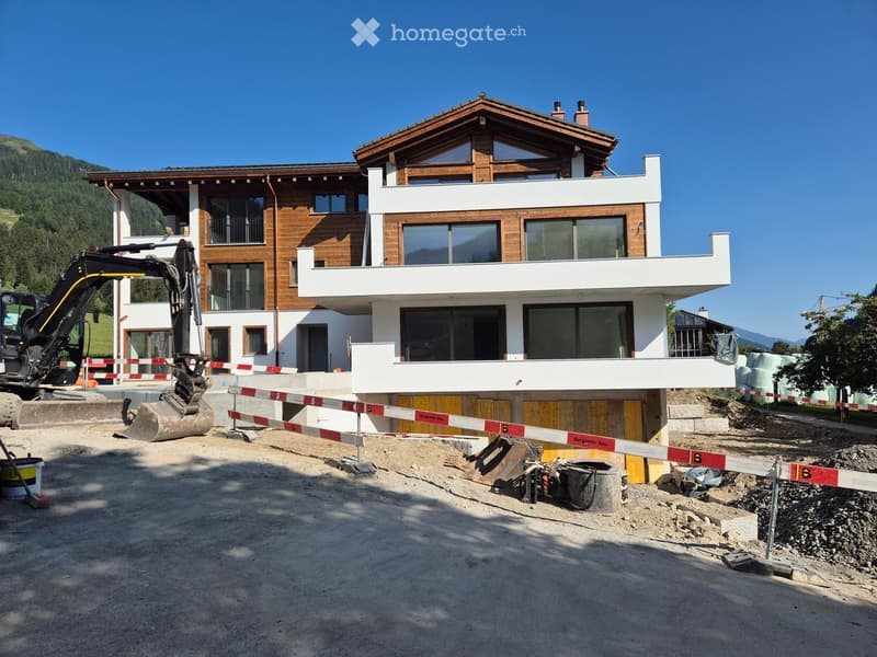 Modern two-story house under construction with wooden and white exterior, large windows, and a raised ground floor. An excavator is visible near the site.