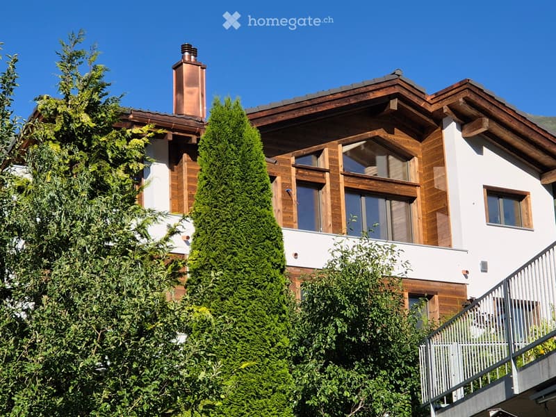 A two-story house with a wooden facade, large windows, and a balcony. The roof is covered with wooden shingles and has a copper chimney. The house is surrounded by lush green trees.