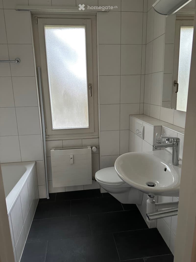 Modern bathroom with a bathtub, toilet, sink, and a radiator. The walls are tiled, and the floor is dark grey.