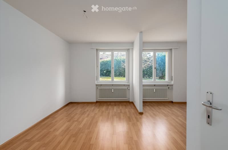 Completely empty room with bare white walls and wooden floor. Two windows with white frames and radiators below them. No furniture or equipment.