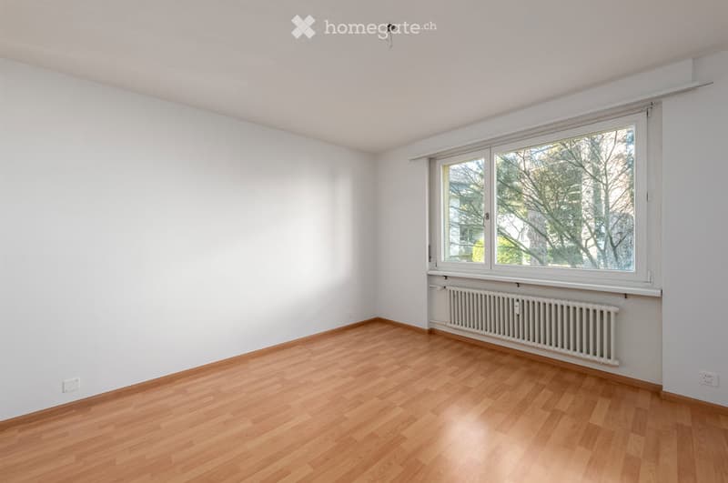 Empty room with light wooden flooring, white walls, and a large window with a radiator below it.