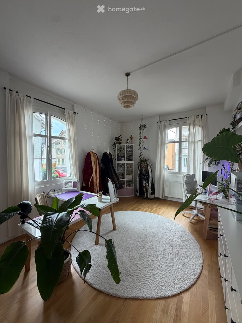 A well-lit room with wooden flooring, large windows, and a round white rug. Contains a table with a plant, a chair, shelves with books and decorative items, and a hanging lamp.