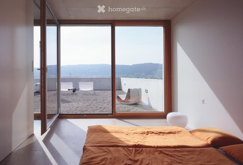 Bright bedroom with large windows offering a scenic view, featuring a bed with orange bedding, a white chair, and a small table.