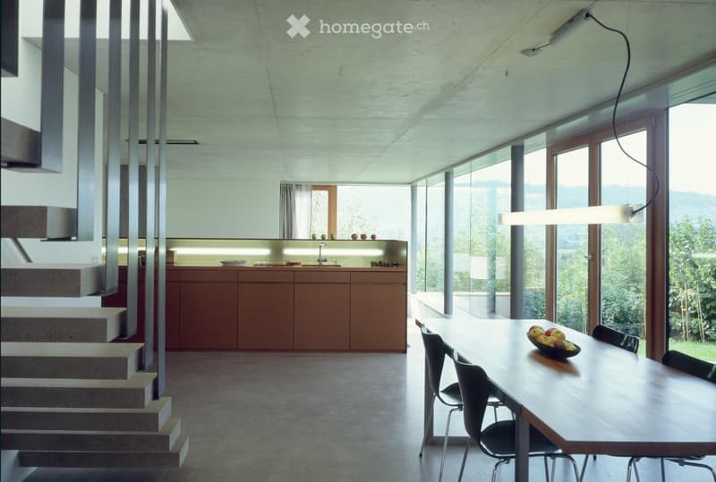 Modern, open-plan kitchen and dining area with large windows, wooden table, chairs, and a sleek kitchen counter with a faucet.