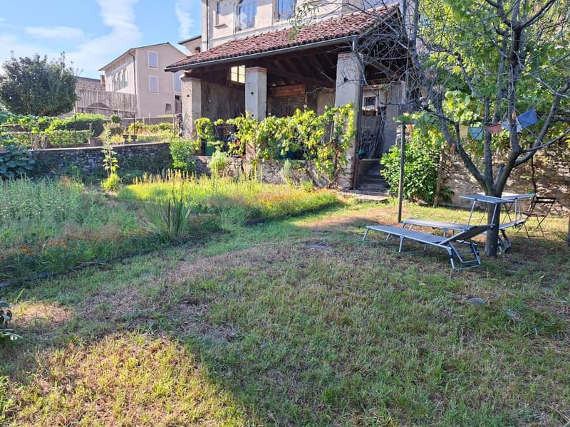 Garden area with a stone wall, grassy lawn, and outdoor furniture including a table and chairs near a tree. A rustic building with a tiled roof is visible in the background.