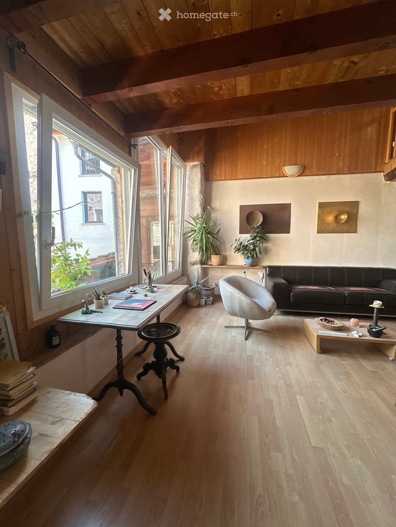 Bright living room with wooden floors, large windows, a black leather sofa, a white armchair, a wooden coffee table, and various decorative items including plants and art pieces.