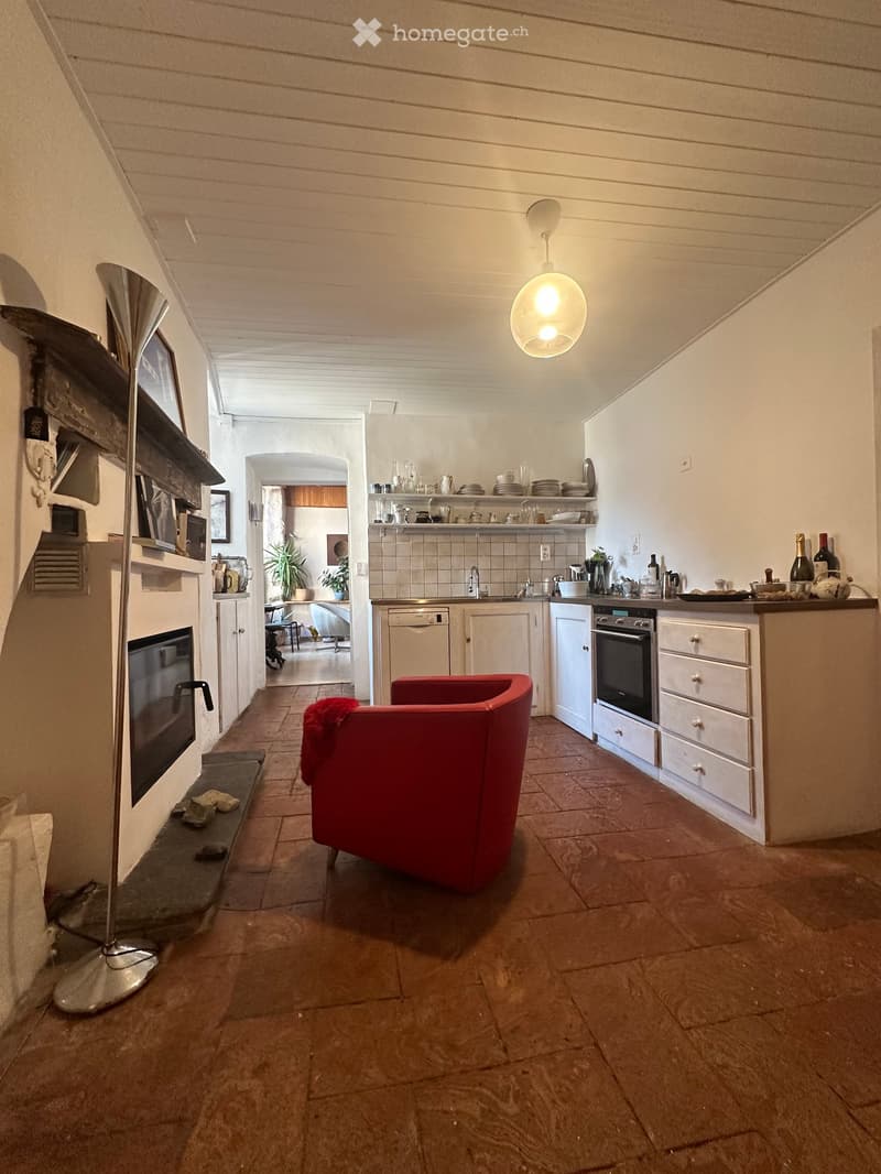 A cozy kitchen area with a red chair, white cabinets, a kitchen island, shelves, and a modern lamp. The space features a tiled floor and a fireplace with a floor lamp nearby.