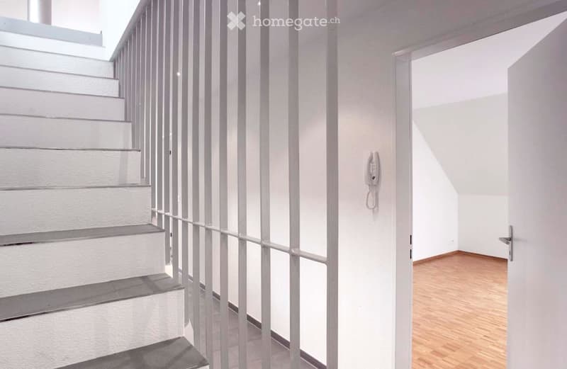 Modern hallway with white walls, wooden flooring, and a staircase with white railings. A telephone is mounted on the wall, and a door leads to another room.