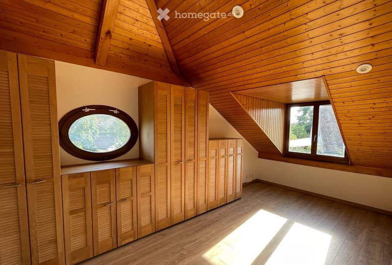 Bright, empty attic room with wooden walls, ceiling, and flooring. Features built-in wooden cabinets, a round window, and a large window with a view of the outside.