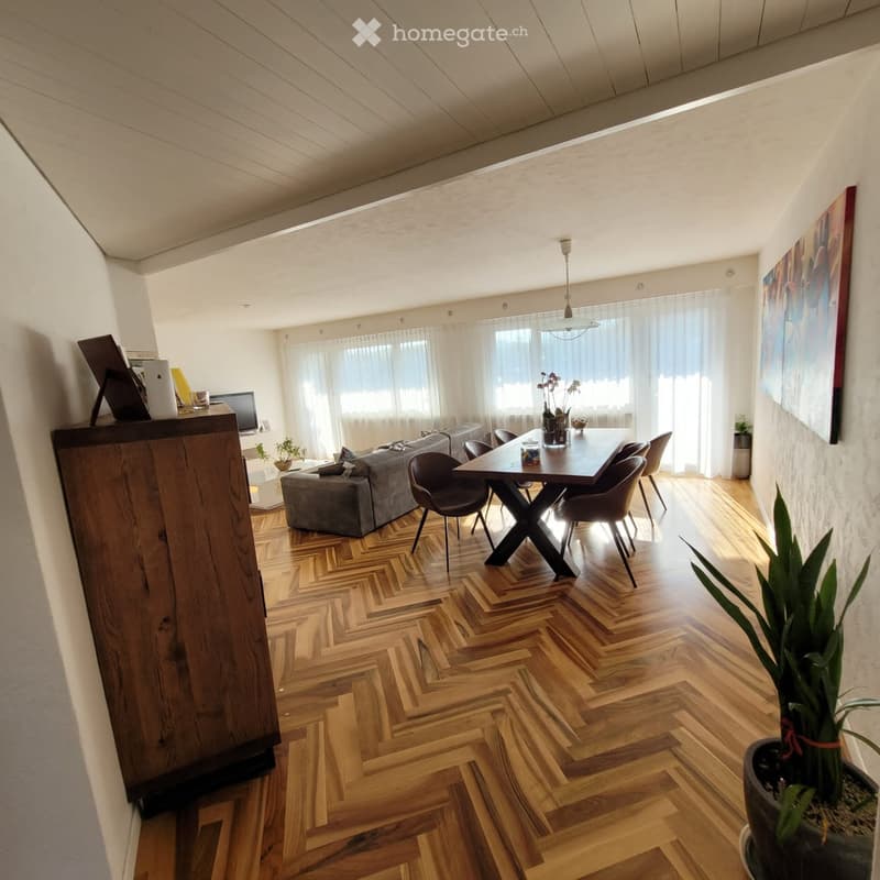 Modern living and dining area with herringbone wooden flooring, large windows with sheer curtains, a grey sofa with ottoman, a wooden dining table with four chairs, and a potted plant near the window.